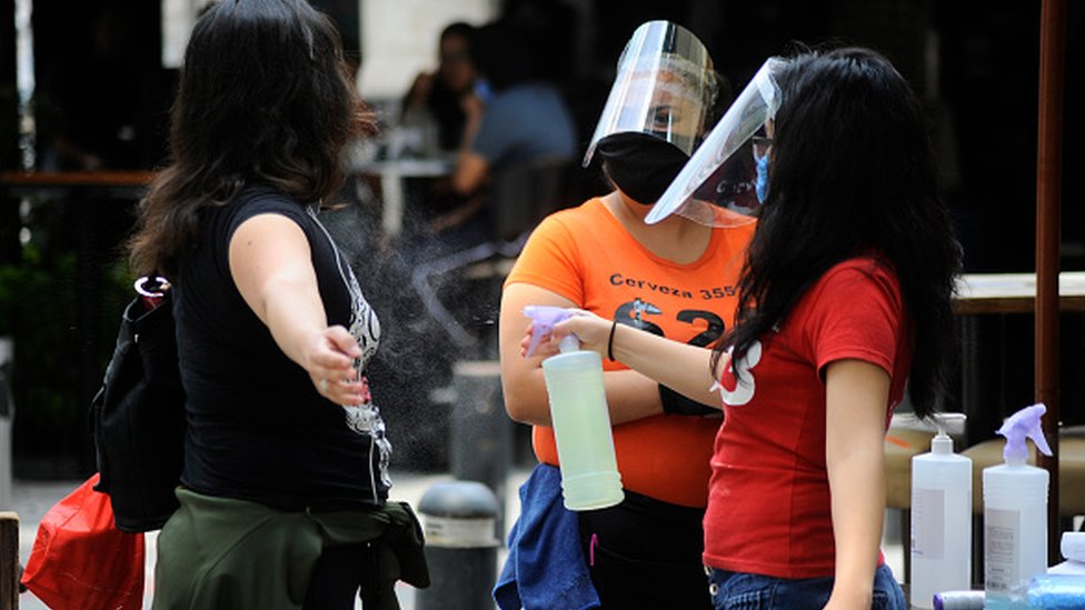 Хозяйка надевает защитную маску, распыляя на человека антибактериальный раствор перед тем, как войти в ресторан Zona rosa в Мехико