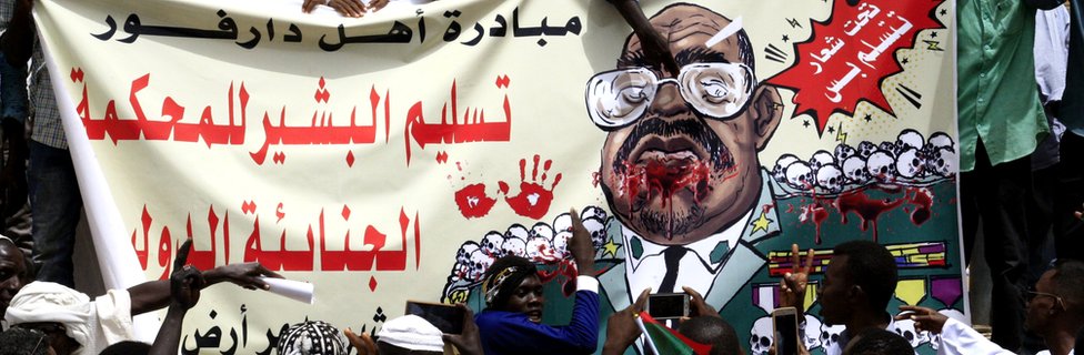 Суданские протестующие держат плакат с изображением свергнутого президента Омара аль-Башира с текстом на арабском языке: «Инициатива народа Дарфура: передача аль-Башира Международному уголовному суду (МУС)» в столице Хартуме, 19 апреля 2019 г.