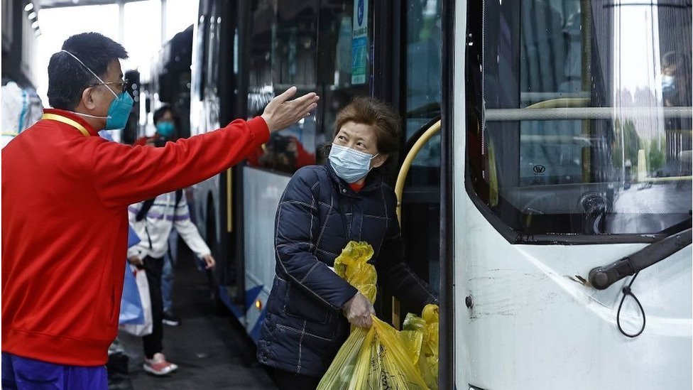 Um oficial usando uma máscara gesticula para uma mulher de meia-idade enquanto ela entra em um ônibus segurando uma grande sacola plástica com pertences