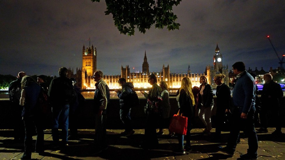 People queue along London's South Bank at night with the Palace of Westminster in the distance