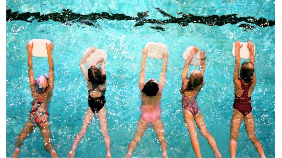 Foto tirada de cima mostra cinco crianças em aula de natação, usando pranchas
