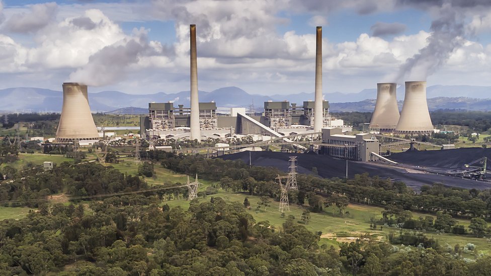 Grey power plant in the middle of green area, with smoke coming from towers into blue sky