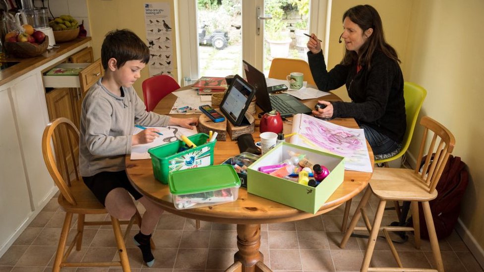 Uma mulher e o filho na mesa de casa; enquanto ela trabalha, ele desenha