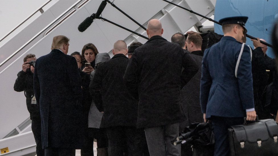 Donald Trump de espaldas siendo entrevistado por periodistas en la pista de un aeropuerto.