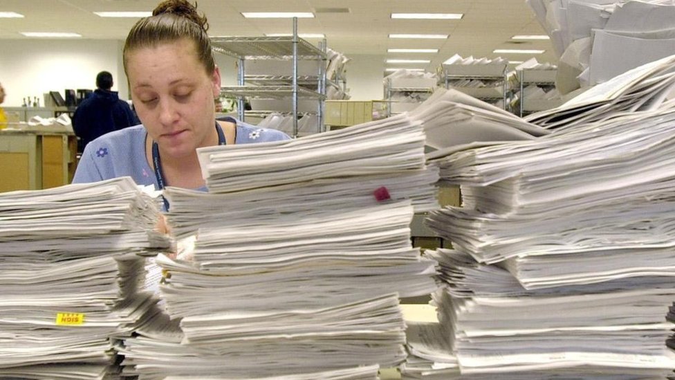 Toma el primer papel de la pila. Dar el primer paso siempre es mejor que no dar ninguno. Una mujer detrás de varias pilas de papeles