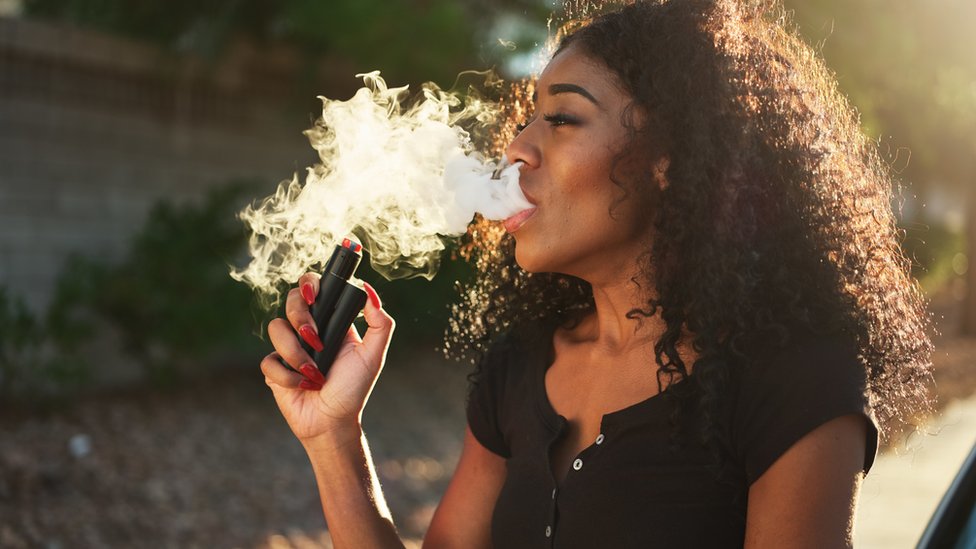 Una mujer, con una camiseta negra, exhala una gran nube de vapor de cigarrillo electrónico, mientras fuma de un vaporizador.
