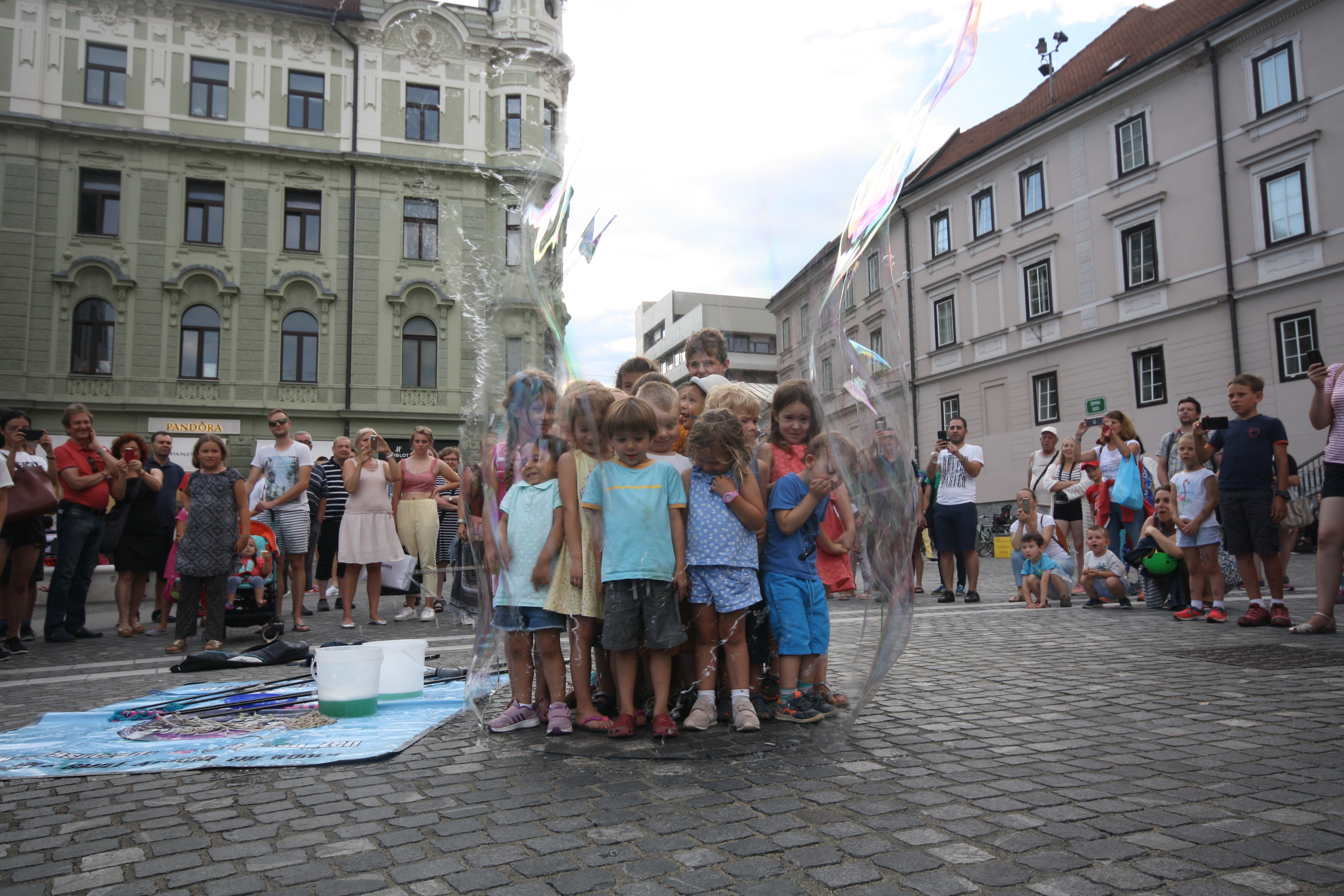 Imagem mostra crianças dentro de uma bolha de sabão observadas por grupo de adultos, em uma praça na Europa
