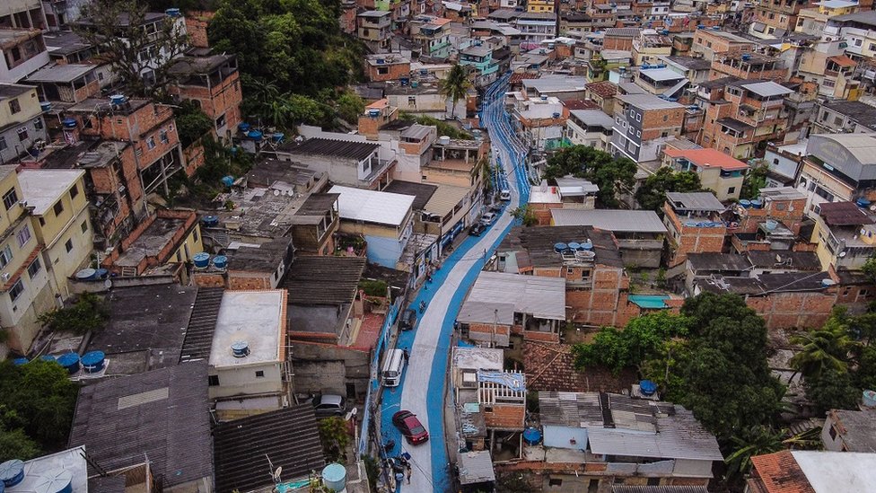 Foto aérea mostra rua pintada de azul e branco em meio às dezenas de casas da favela