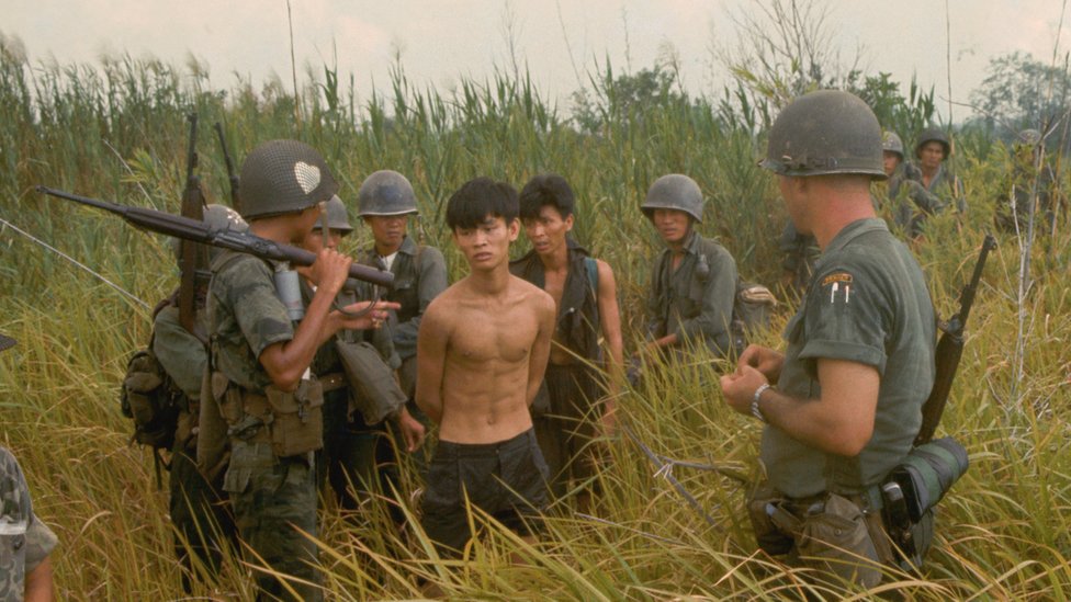 Sekelompok tentara tentara Vietnam Selatan dan seorang tentara Amerika dengan dua tersangka Vietcong yang ditangkap.