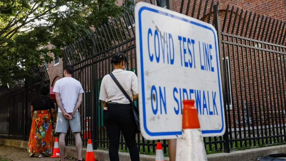 Três pessoas na rua, em fila, perto de placa dizendo em inglês: teste para covid