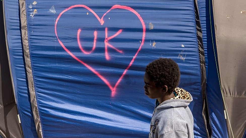 Мигрант идет рядом с палаткой с инициалами Великобритании в лагере для мигрантов и беженцев в Кале