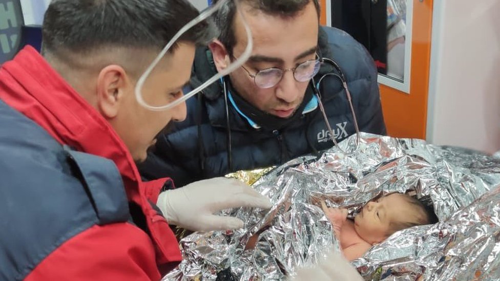 Yagiz, un bebé de apenas 17 días, envuelto en una manta térmica es atendido por dos hombres.