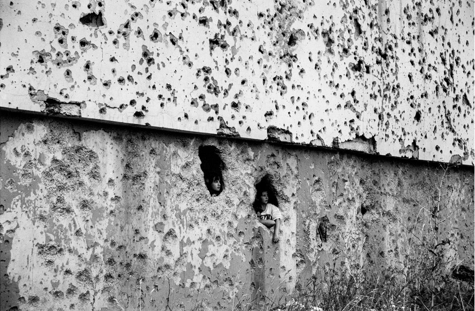 Дети выглядывают из дыр в стене, покрытой пулевыми отверстиями
