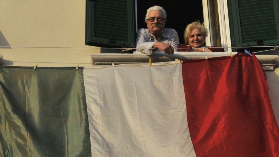 Una pareja mayor en un balcón con una bandera italiana.