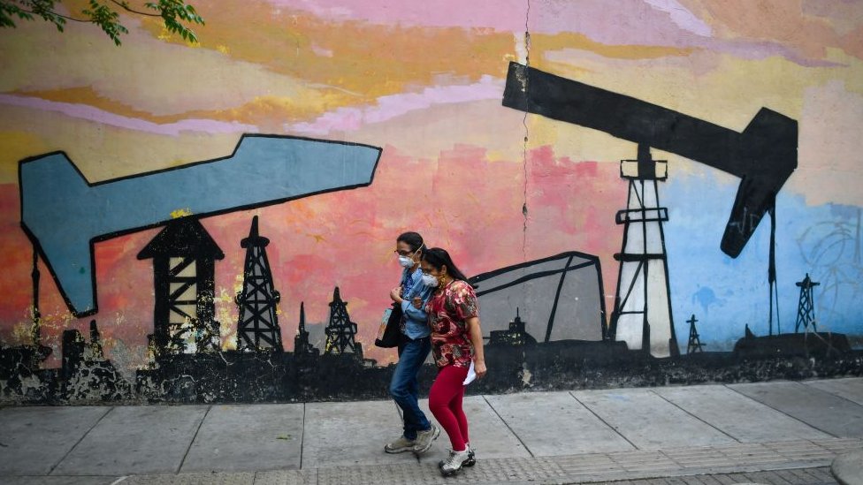 Dos mujeres caminan por la calle en Venezuela junto a un mural en el que hay pintados balancines de petróleo.