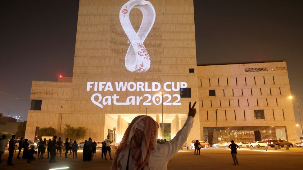 Homem com lenço usado comumente na cultura árabe em frente a um prédio com a sinalização da Copa do Mundo