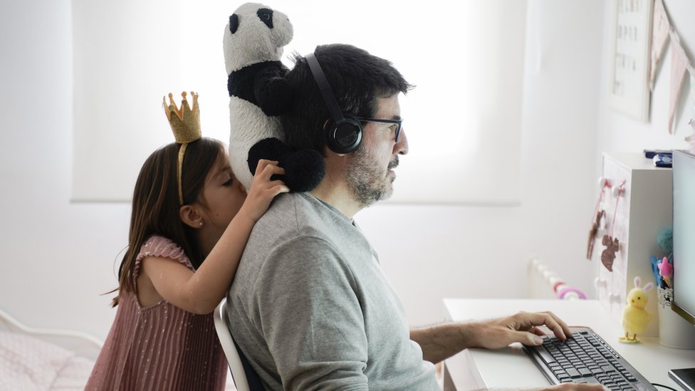 Homem usa o computador, enquanto menina coloca um ursinho de pelúcia em seu pescoço