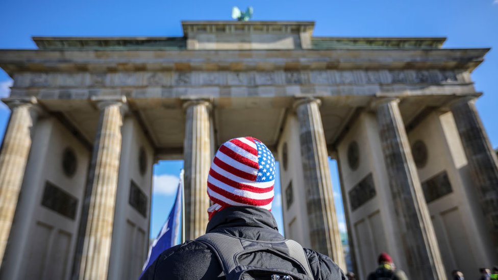 A man in a US flag hat stands in front of Brandenburg Gate