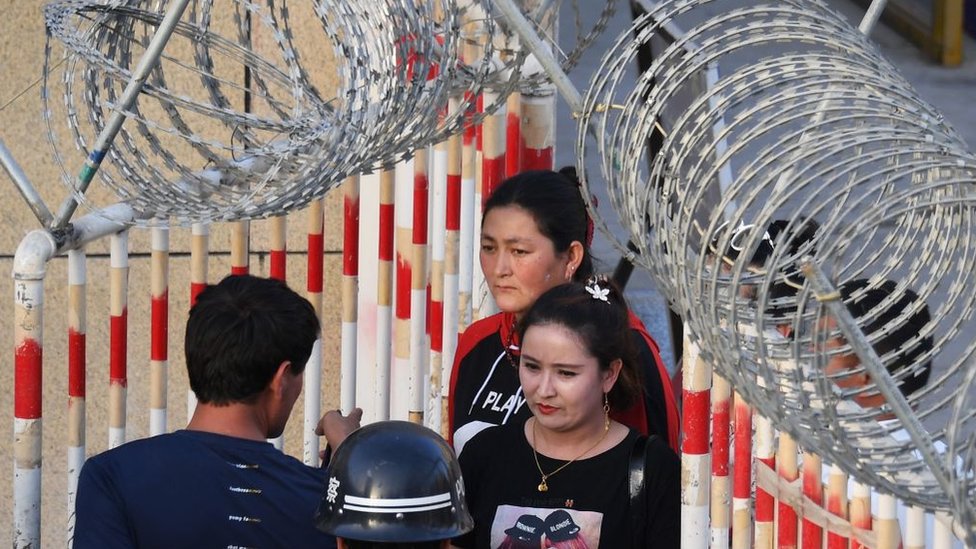 A file photo from 2019 showing Uygher women going through the entrance to a bazaar in Hotan, Xinjiang, past police officers and a gateway covered in razor wire