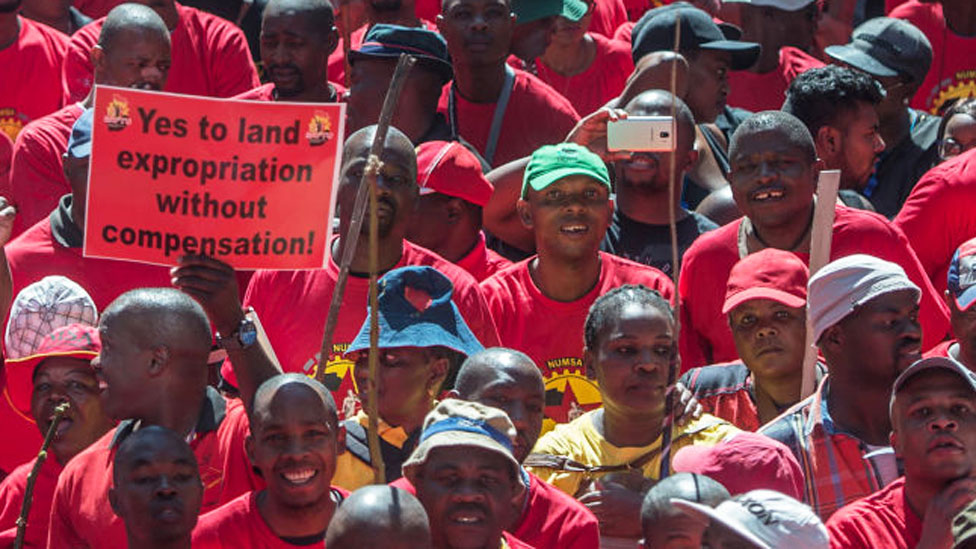 Демонстрант в Южной Африке держит плакат с надписью «Да экспроприации земли без компенсации», когда тысячи рабочих принимают участие в национальной забастовке, объявленной вторым по величине профсоюзом страны - апрель 2018 г.