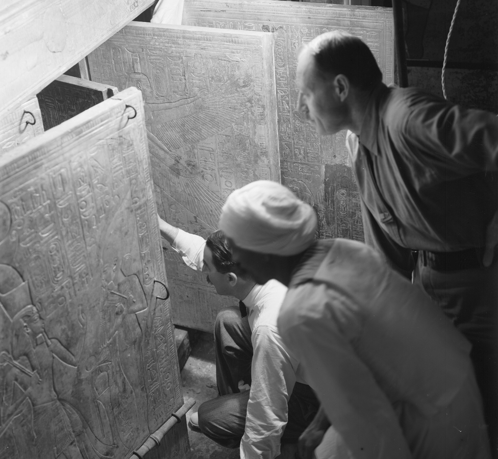 Foto em preto e branco mostra dois homens brancos com bigode e um trabalhador egípcio abrindo portas gravadas com hieróglifos