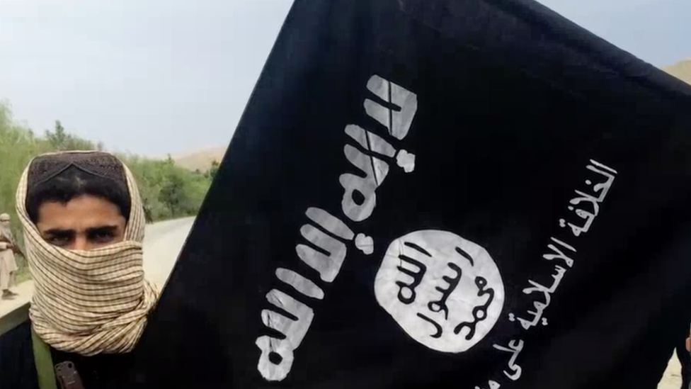 Скриншот видео ИГ, на котором мужчина в шарфе стоит рядом с флагом ИГ
