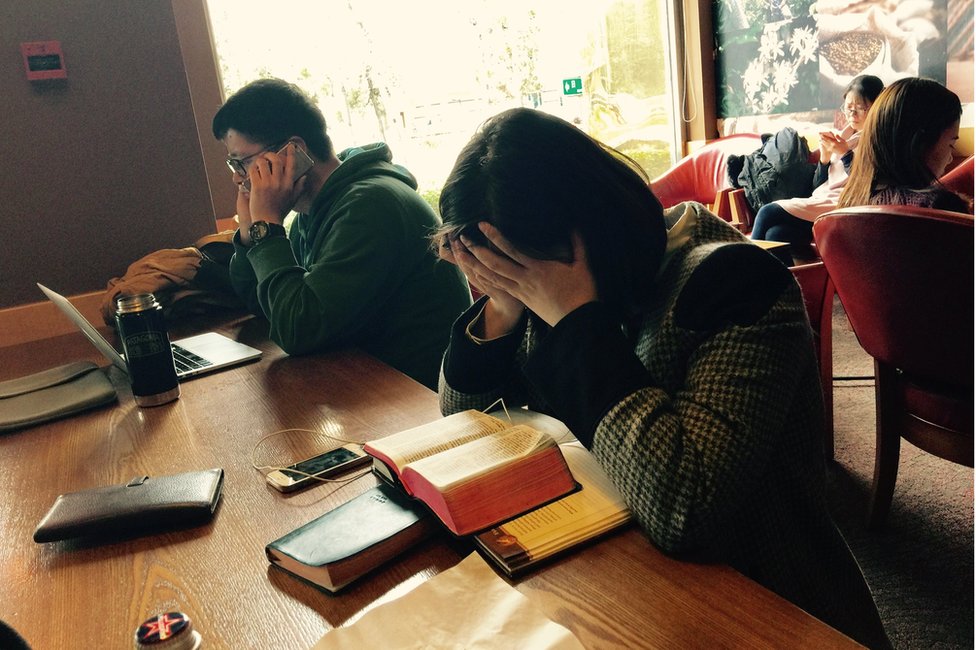 Кафе в Китае, где женщина держит голову руками перед Библией, а мужчина разговаривает по мобильному телефону перед ноутбуком.