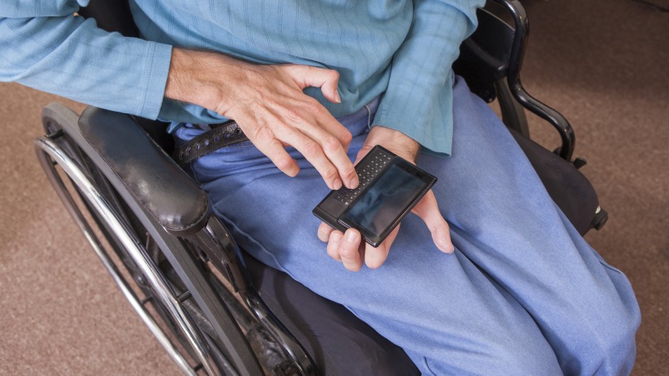 Homem branco sentado em cadeira de rodas usando um aparelho para auxiliá-lo a falar