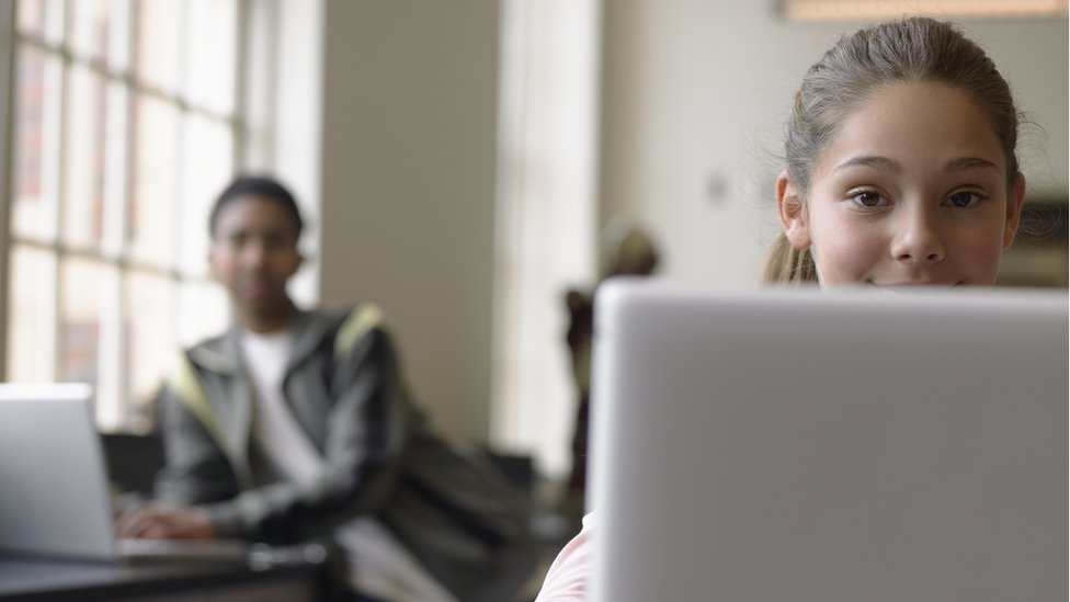 A young girl sits at a laptop while a male student, blurred by the camera, sits behind