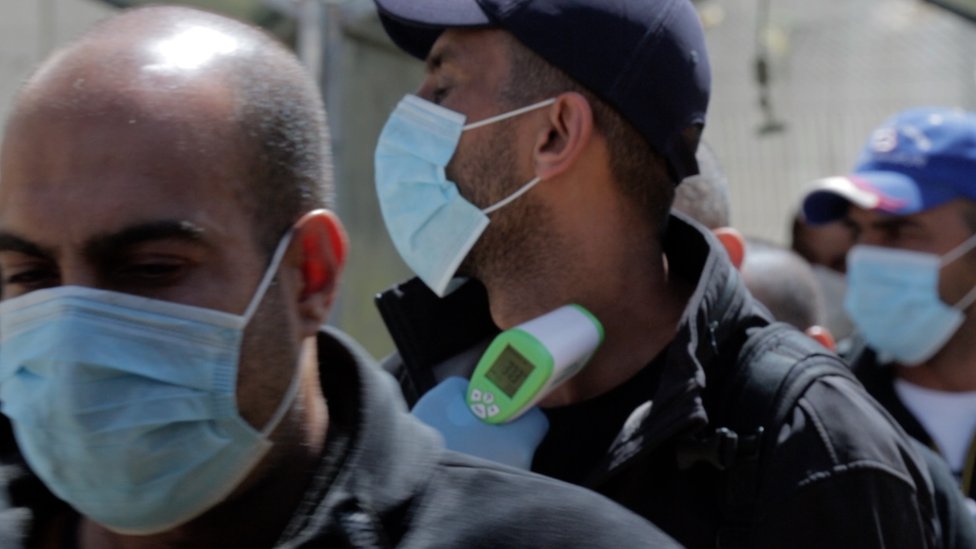 Палестинские медицинские работники проверяют температуру рабочих, переходящих через контрольно-пропускной пункт Таркумия, недалеко от города Хеврон на Западном берегу реки Иордан