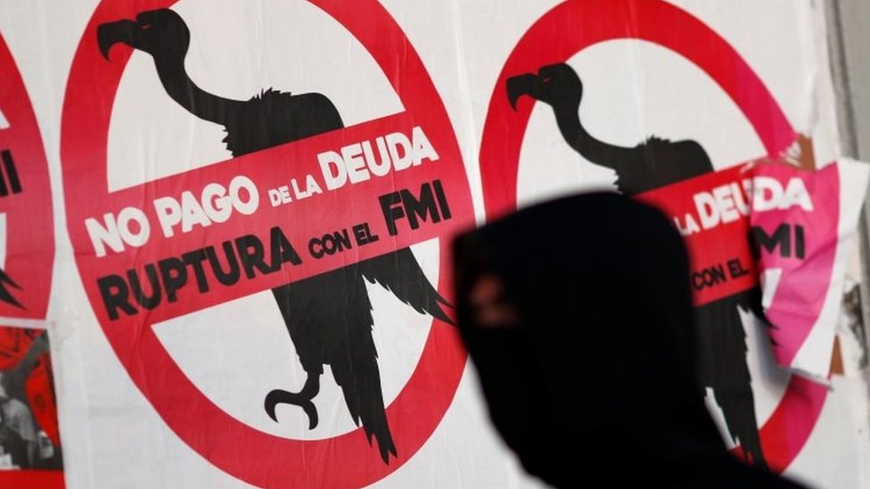 Пешеход в маске в качестве профилактики коронавирусной болезни (COVID-19) проходит мимо плакатов на улице с надписью «Нет выплате долга. Разрыв с МВФ»