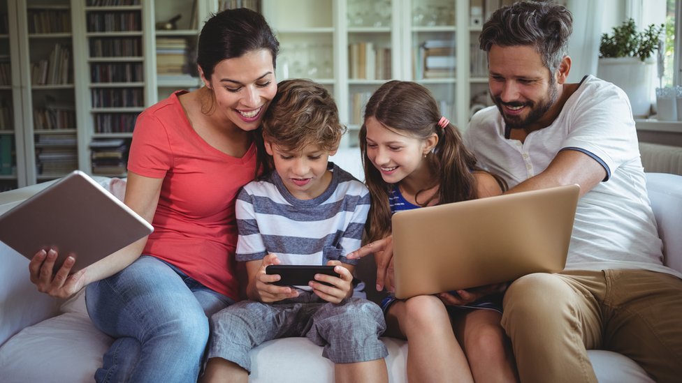 أسرة من أب وأم وابن وابنة يتابعون أجهزة إلكترونية