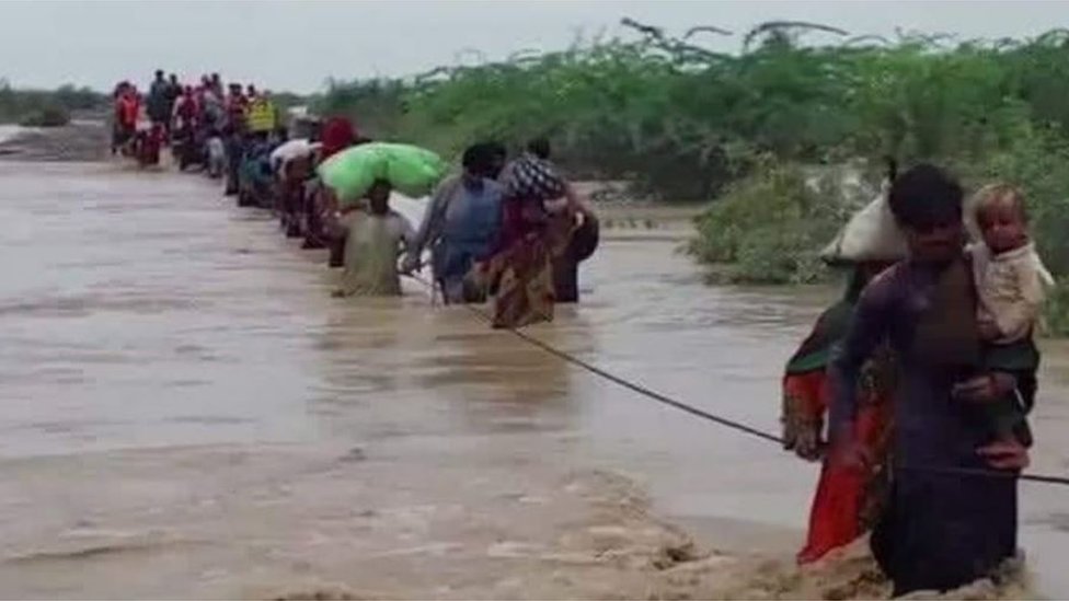 Moradores de vilarejos atravessam as margens de um rio que transbordou segurando uma corda de segurança