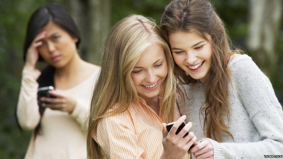 Две девушки улыбаются и смотрят в телефон, а другая выглядит грустной