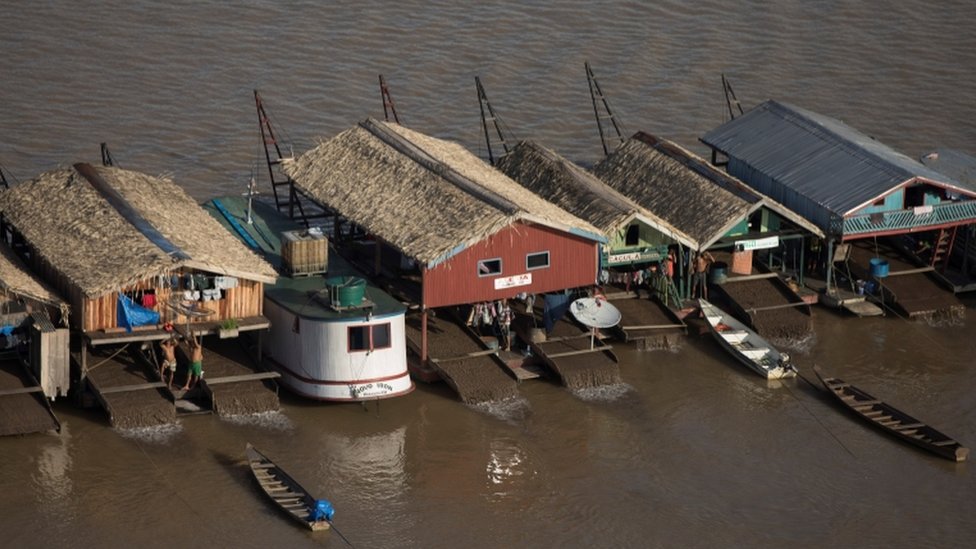 Foto aproximada mostra casas e barcos instalados no meio do rio