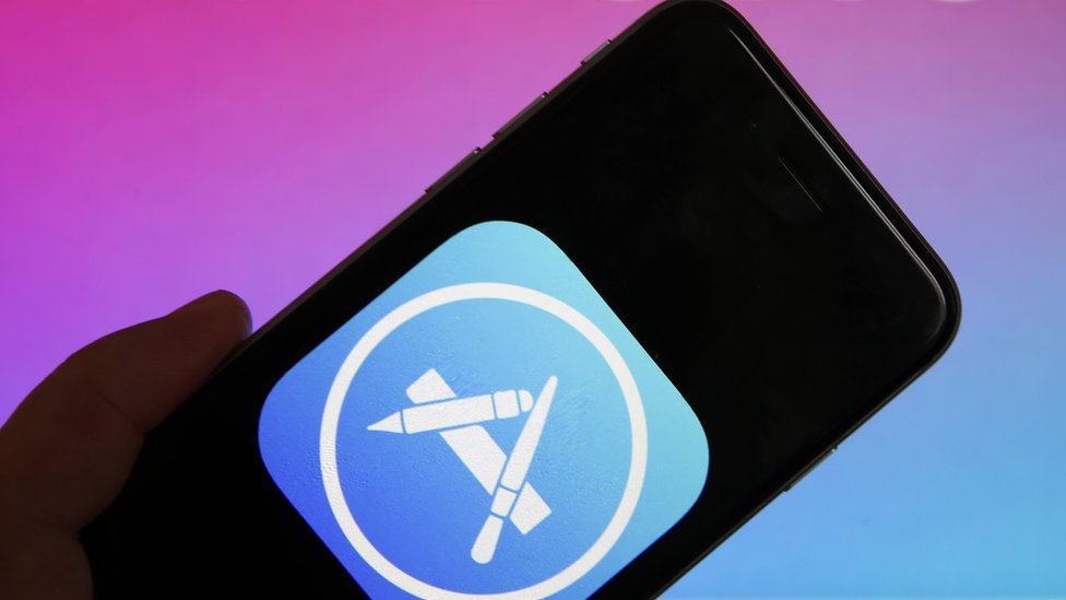 A photo illustration shows the Apple App Store logo displayed on an iPhone held up against a blue-pink gradient background