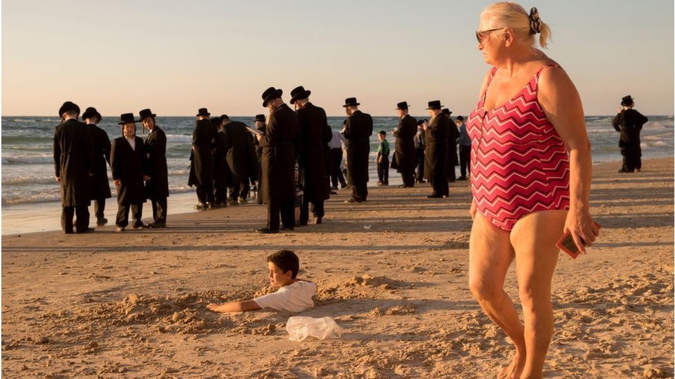A woman in a bathing suit watches a group of Orthodox Jews on a beach near Tel Aviv