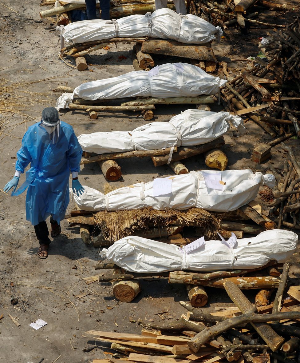 A health worker wearing personal protective equipment (PPE) walks past a row of wrapped bodies of Covid victims lying on prepared funeral pyres in New Delhi, India