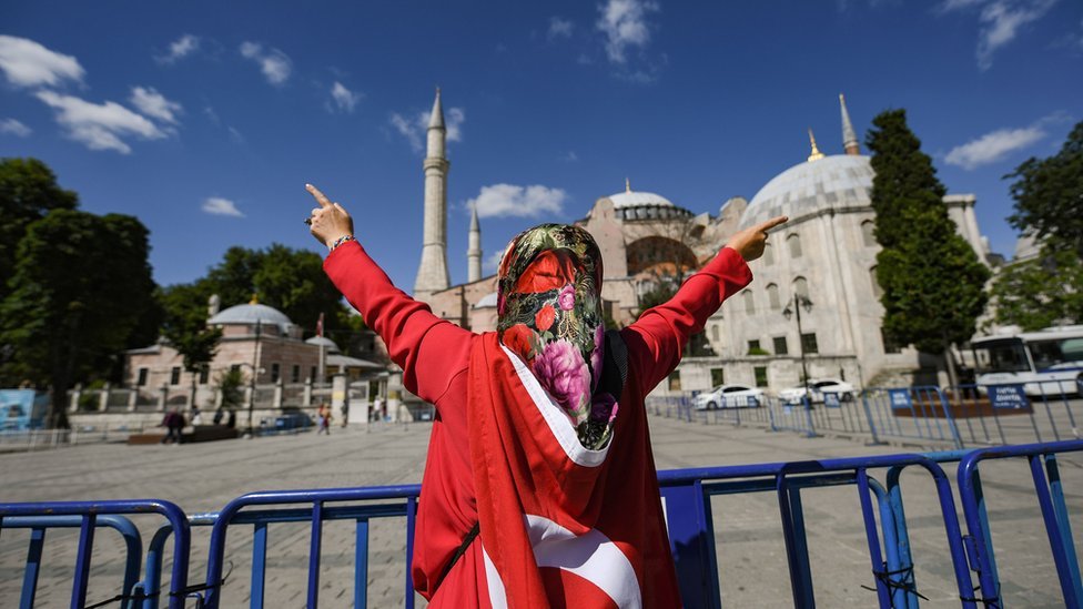Женщина, одетая в турецкий национальный флаг, жестикулирует у музея Святой Софии 10 июля 2020 года в Стамбуле, когда люди собираются, чтобы отпраздновать после того, как высший турецкий суд отменил статус собора Святой Софии шестого века как музея, расчистив путь превратится обратно в мечеть