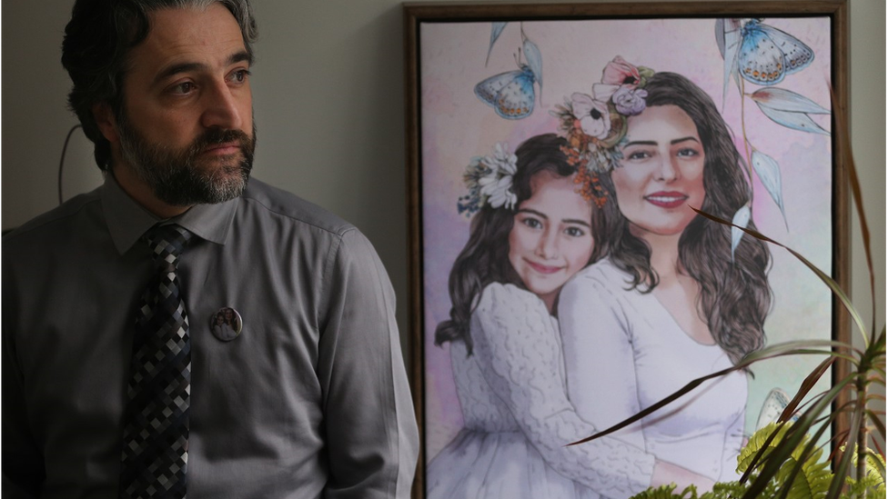 Hamed Esmaeilion standing in front of a painting of his wife and daughter and also wearing a pin with the same image
