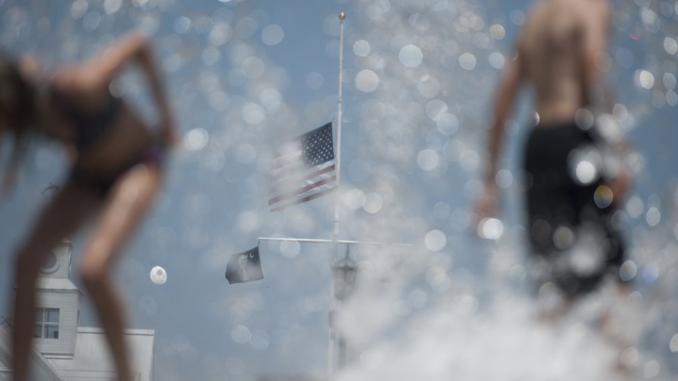 2015-06-21. Флаг развевался наполовину, когда дети играли в фонтане в парке Уотерфронт в Чарльстоне. Колм О'Моллой для BBC News.