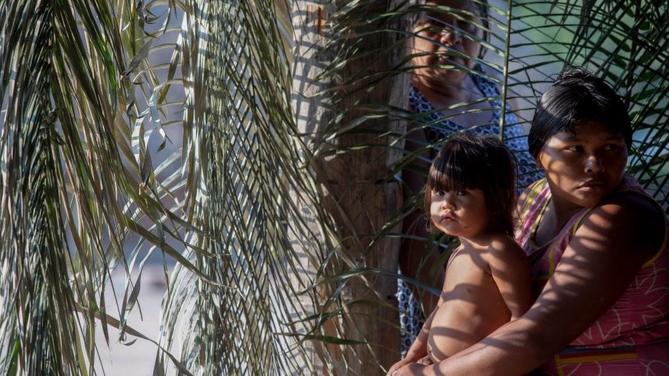 Mulher indígena de pé ao lado de outra mulher e uma criança, também indígenas, cercados por folhas de palmeira
