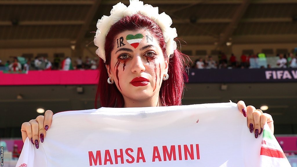 A female fan in the crowd for Wales v Iran holds up a football shirt with "Mahsa Amini - 22" printed on it