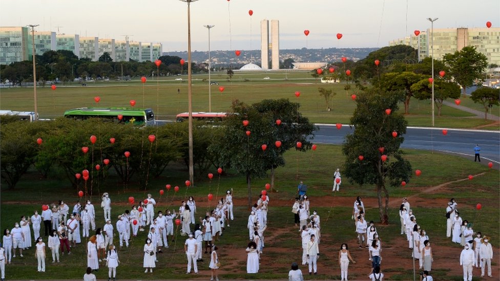 Em demonstração em área aberta de Brasília, diversas pessoas em filas vestidas de branco soltam balões vermelhos em homenagem a vítimas da covid-19