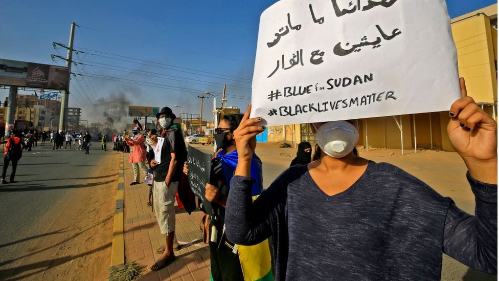 Протестующий из Судана держит табличку с надписью на арабском языке «Наши мученики не мертвы, они живы с революционерами» вместе с английскими лозунгами «#BLUEforSUDAN» и «#BLACKLIVESMATTER», когда демонстранты отмечают первую годовщину рейда на антиправительственная сидячая забастовка и некоторые демонстрации в поддержку протестующих в США в связи со смертью Джорджа Флойда в районе Эр-Рияд на востоке столицы Хартума 3 июня 2020 г.