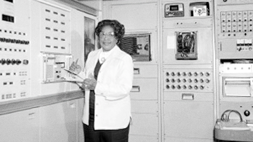 Мэри Джексон держит планшет перед большим компьютером