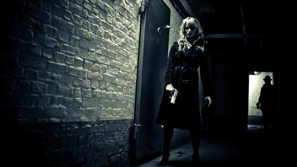 Una mujer espía con un arma en un corredor observada por un hombre a la sombra desde el umbral de una puerta