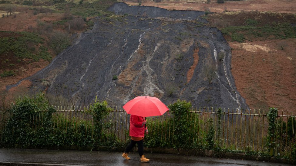 Женщина с зонтиком идет мимо оползня на угольной оконечности в Тайлорстауне, Ронда Кинон Таф