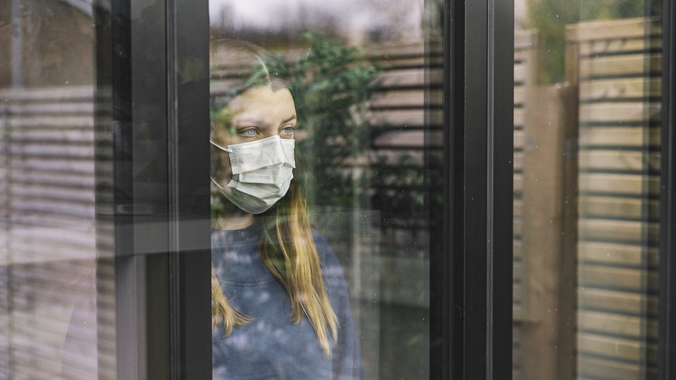Una mujer con mascarilla detrás de una ventana.