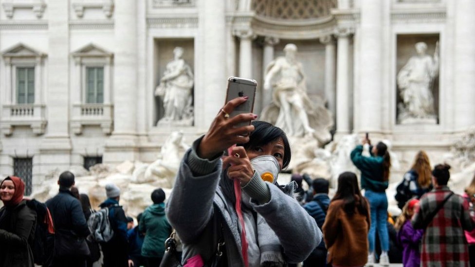 Китайский турист у фонтана Треви в Риме делает селфи в маске